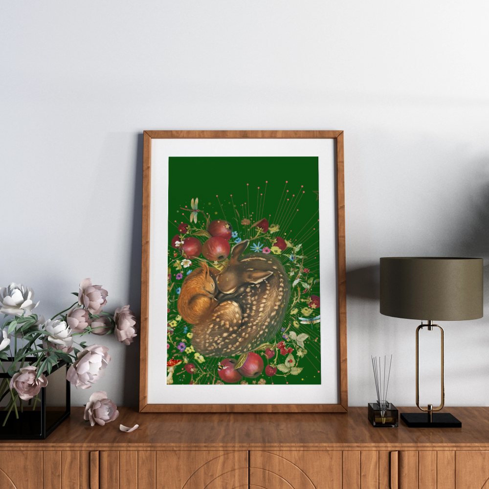 A framed art print featuring a whimsical forest illustration of a deer and squirrel among red apples and wildflowers on a dark green background, styled on a wooden console table next to a lamp and flowers.